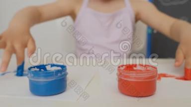 一个小可爱的女孩坐在桌子旁，用明亮的手指<strong>颜料画</strong>纸，把她的手指浸在颜料罐里。